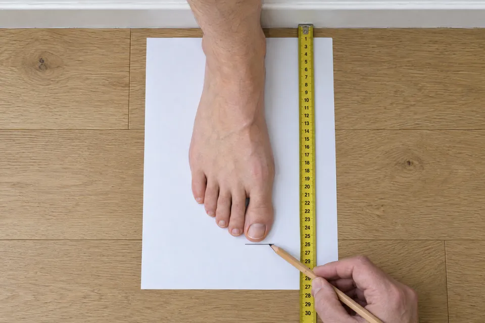 איך מודדים אורך כף רגל. עקב צמוד לקיר, דף נייר מתחת לרגל, סימן בקצה הבוהן הארוכה ביותר, והמדידה נלקחת מהקיר עד הסימן בסנטימטרים.